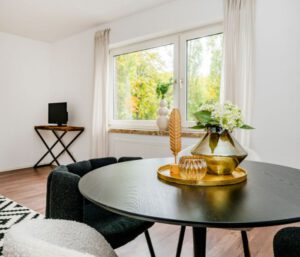 Luxuriöser Essbereich mit runder schwarzer Tischplatte, goldfarbenem Deko-Set und großer Fensterfront, hochwertiges Interieur bei Wealth-Living Immobilien.